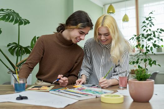 Teenagers painting with watercolors. Couple of guy and girl sitting at table with paints, brushes, drawings. Happy teens talking laughing creative. Creativity, hobby, leisure concept