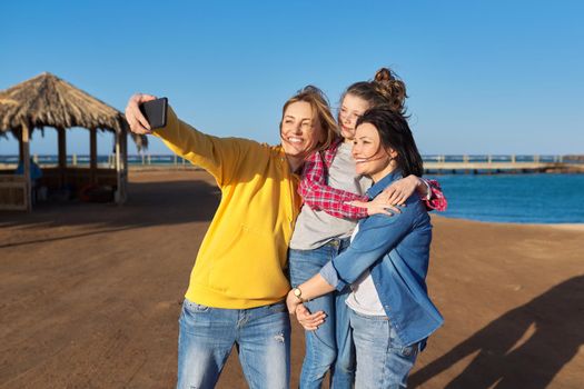 Happy smiling two middle aged women and girl child taking selfie photo together on smartphone, sea beach sky background. Vacation, travel, happiness, joy, family, lifestyles, people concept