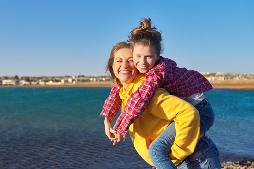 Happy mom and daughter child on seashore, relaxing on sandy beach, autumn winter spring season, copy space, blue sky background. Family vacation, travel, parent child relationship, happiness, joy