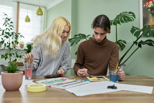 Teenagers painting with watercolors. Couple of guy and girl sitting at table with paints, brushes, drawings. Happy teens talking laughing creative. Creativity, hobby, leisure concept