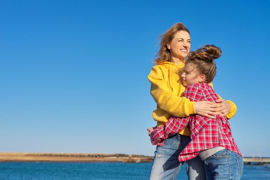 Happy mom and daughter child on seashore, relaxing on sandy beach, autumn winter spring season, copy space, blue sky background. Family vacation, travel, parent child relationship, happiness, joy