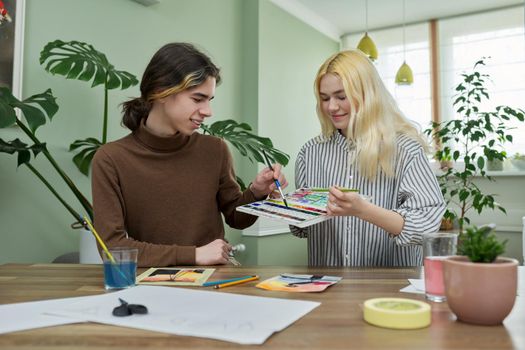 Teenagers painting with watercolors. Couple of guy and girl sitting at table with paints, brushes, drawings. Happy teens talking laughing creative. Creativity, hobby, leisure concept