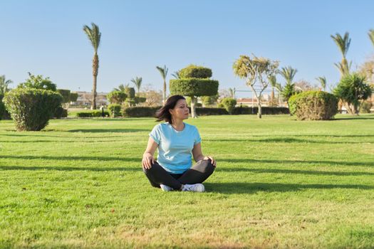 Middle aged woman sitting on grass in lotus position and meditating with closed eyes, sunny morning on a summer day. Meditation, relaxation, healthy lifestyle and body, mental health, people 40s