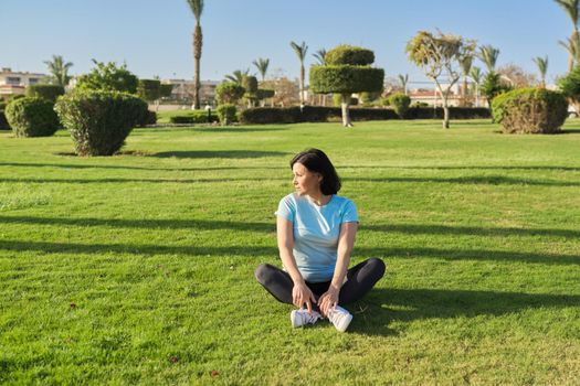 Middle-aged woman doing outdoor sports, 40s female sitting on grass on sunny summer morning. Healthy lifestyle, healthy body, mature people concept