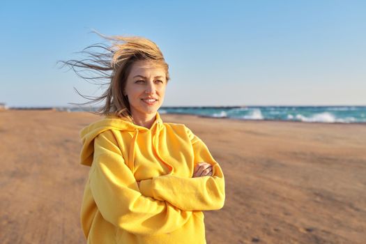 Outdoor portrait of smiling happy blonde woman 45 years old looking at camera with crossed arms. Sunny day on the sea coast, beach blue sky background
