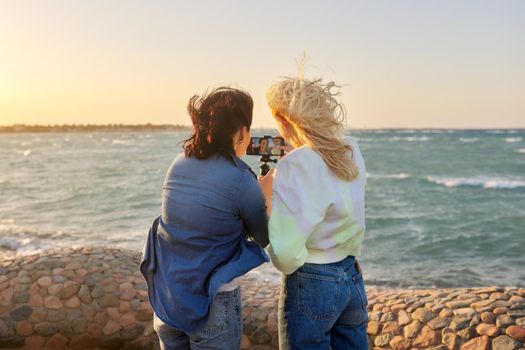 Sea vacation, family mother and teenage daughter together on the sea coast at sunset. Females look at smartphone screen, make an online video call, season is tropical autumn winter spring