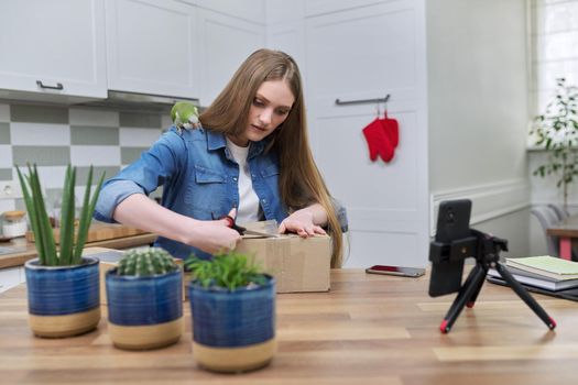 Woman recording video on smartphone moment of unpacking cardboard box. Showing purchases ordered on Internet, product quality reviews with followers, blog content, feedback. Pet parrot helping unpack