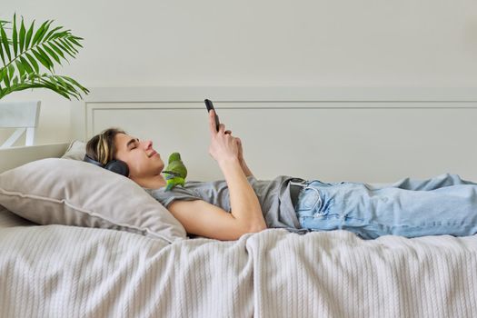 Male teenager lying in home bed with pet green parrot Quaker, wearing headphones and smartphone in his hands. Lifestyle, leisure, relax, teenagers, love for pets, technology concept