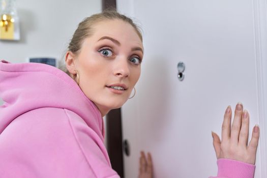 Young woman looking through the peephole of front door in apartment. Emotion of surprise on female's face