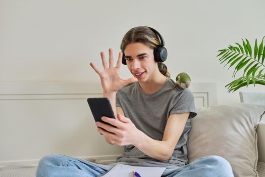Emotional talking male teenager in headphones looking at smartphone screen using video call. Bird pet green quaker parrot on the owners shoulder, home room sofa bed background