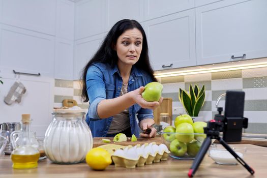 Woman cooking apple pie at home in the kitchen, talking online using a video call on a smartphone, showing green apples on webcam