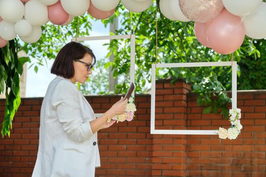 Decorating the garden with balloons for party, ceremony. Woman professional event organizer with digital tablet, creative work, event company, business female small business owner organizing holiday