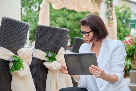 Organization of weddings, parties, event decoration. Business woman professional decorator with digital tablet working outdoors decorating ceremony. Female against the background of decorated garden chairs