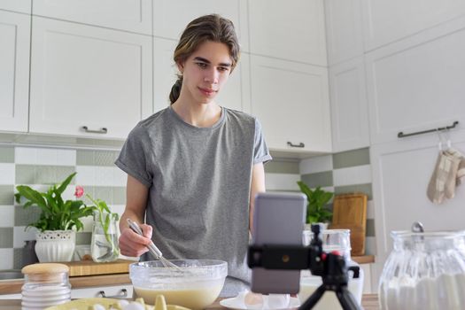 Male teenager learns to cook pancakes, watching video recipe on smartphone, in home kitchen. Cooking home food, culinary hobby, learning cooking class, teenagers, people concept