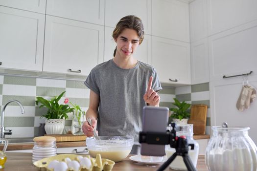 Male teenager cooking pancakes talking on video communication on smartphone, home kitchen interior background. Guy learns to cook, talks with family, shows recipe. Online technology, people, lifestyle concept