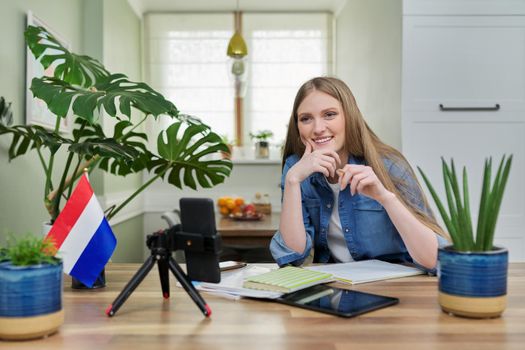 Young female university student sitting at home studying online, looking at smartphone webcam. On table flag of Netherlands, education in Holland, e-education, e-learning, technology, knowledge
