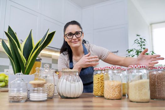 Storing food in kitchen, woman housewife in an apron with jars and containers of cereals, food, pasta, talking and looking at camera. Female blogging about organizing space in kitchen