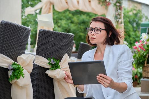 Organization of weddings, parties, event decoration. Business woman professional decorator with digital tablet working outdoors decorating ceremony. Female against the background of decorated garden chairs