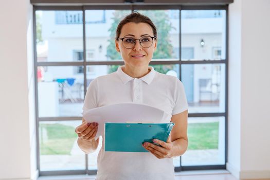 Business woman portrait with clipboard in hand. Middle-aged female wearing white T-shirt glasses, inside office hall. People, workers, profession, staff, occupation, administration concept