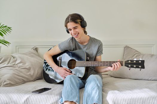 Male teenager in headphones sitting at home on bed with acoustic guitar and smartphone. Hipster guy using smartphone to learn music, study songs, record his music. Creativity, hobby, teens concept