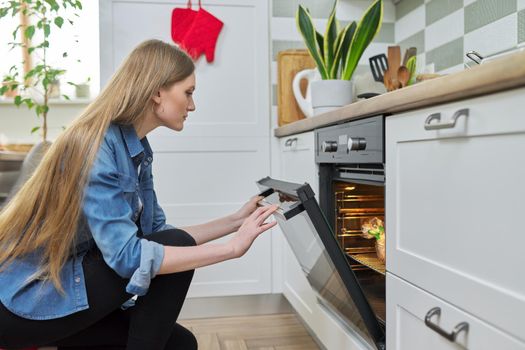 Cooking at home, roasting meat in oven, young woman putting marinated chicken in baking bag in oven, kitchen interior background