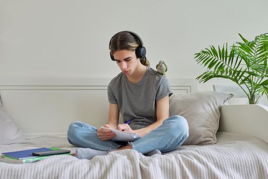 Male teenager with pet green parrot quaker on his shoulder, guy student sitting in headphones with textbooks notebooks books at home on bed