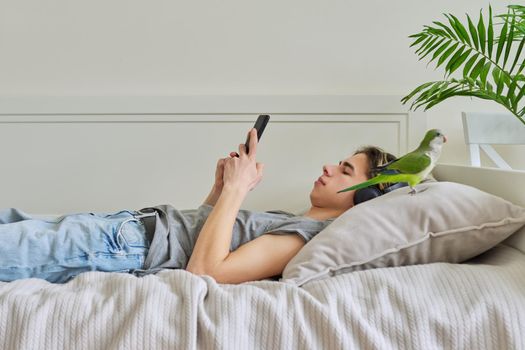 Male teenager lying in home bed with pet green parrot Quaker, wearing headphones and smartphone in his hands. Lifestyle, leisure, relax, teenagers, love for pets, technology concept