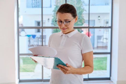 Business woman portrait with clipboard in hand. Middle-aged female wearing white T-shirt glasses, inside office hall. People, workers, profession, staff, occupation, administration concept