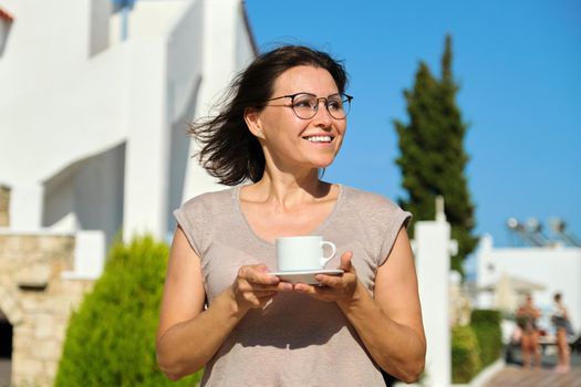 Happy smiling middle-aged female with cup of coffee walking outdoors in the hotel complex, sunny summer day. Vacation, travel, people, tourism