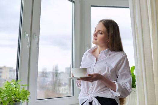 Young beautiful woman with cup of tea at home near window, blonde with long hair in white shirt looking out window, spring winter season, copy space