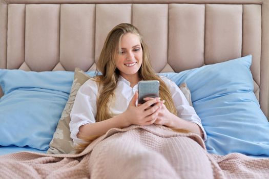 Young beautiful smiling blonde with smartphone lying in bed at home. Successful happy woman looking at the screen, texting, reading, rejoicing