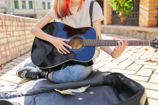 Creative trending female teenager musician playing acoustic guitar sitting on city street sidewalk with guitar case and cash