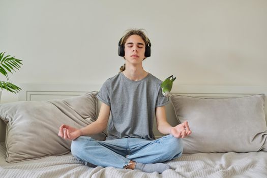 Male teenager sitting in lotus position at home on bed relaxing meditating with closed eyes wearing headphones, bird pet green parrot on shoulder