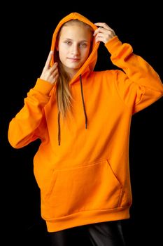 Shot of beautiful stylish teenage girl. Portrait of attractive teenager wearing warm orange hoodie putting the hood on. Beautiful brown eyed girl posing against black background