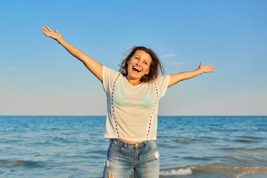 Happy smiling mature woman on the sea beach, positive female with arms raised up. Happiness, joy, relaxation, emotions, middle-aged people