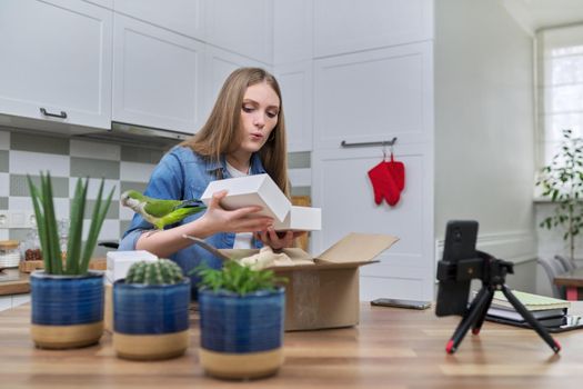 Woman recording video on smartphone moment of unpacking cardboard box. Showing purchases ordered on Internet, product quality reviews with followers, blog content, feedback. Pet parrot helping unpack