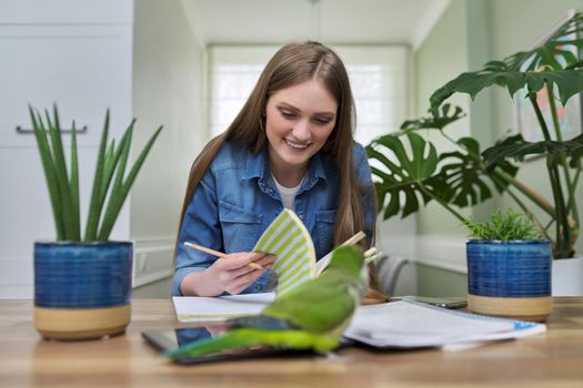 Young female university student studying online using video call, sitting at home at table with pet parrot in front of camera webcam, talking. Technology in education, distance teaching, e-education