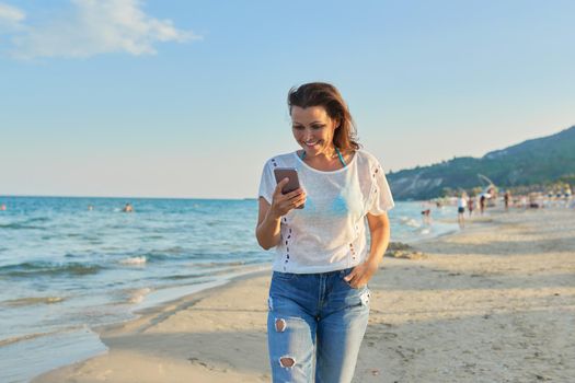 Middle-aged woman walking along beach with smartphone, mature female in jeans white t-shirt resting on sea beach, sunset landscape background. Vacation, weekend by sea, relaxation
