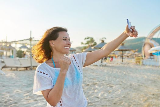 Happy middle-aged woman talking on smartphone using video call, female on sandy sea beach. Summer, vacation, weekend in nature, people of mature age