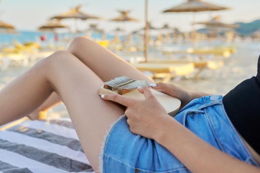 Young woman holding play thumb piano or kalimba musical instrument, female playing music with her hands, relaxing enjoying music and nature on sea beach, sitting in sun lounger at sunset