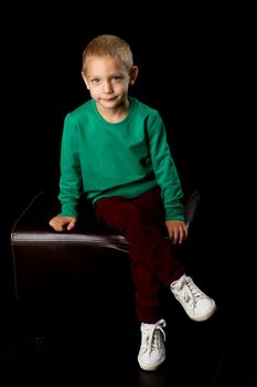 Cute boy posing in front of the camera in the studio, charming boy in a green sweater sits and looks, portrait of a happy blond child, isolated on black background