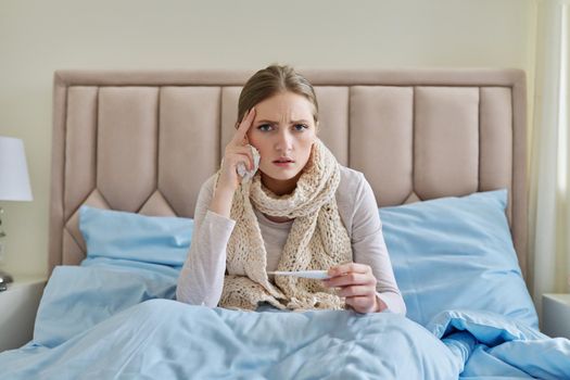 Sick young woman with thermometer in her hands, sitting at home in bed. Upset, unhappy, sad, restless female, high fever, viral symptoms, cold season