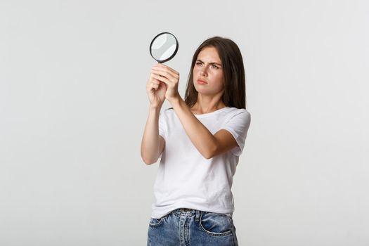 Serious-looking attractive young woman searching for something, looking through magnifying glass, white background.