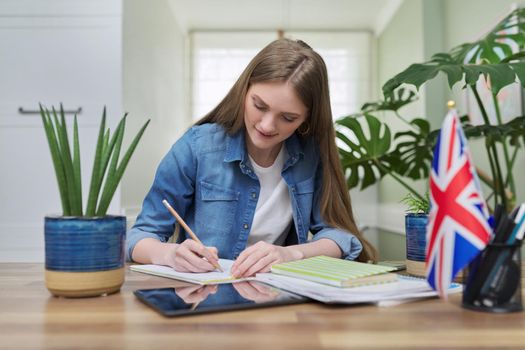 UK learning English online. Female student remotely looking at webcam taking private lessons sitting at home in kitchen, England flag background