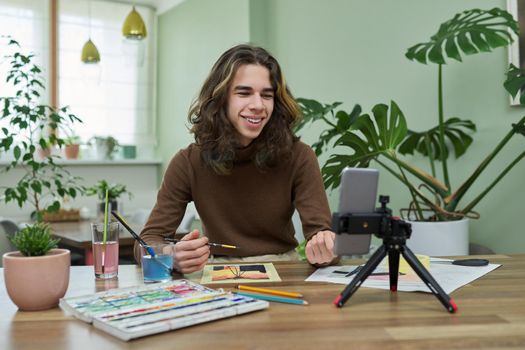 Online learning, drawing, e-education. Teenage guy drawing with watercolors, watching video class on smartphone, sitting at table at home. Creativity, technology, hobby, teens concept