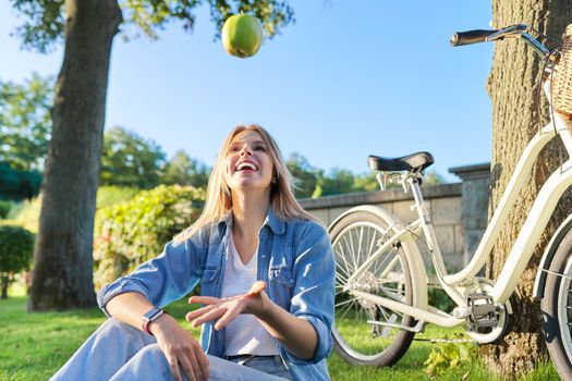 Happy smiling young woman with green apple above her head in air, sitting in park on grass, bike background. Relaxation, healthy active lifestyle, healthy food, happiness and joy
