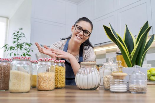 Storing food in kitchen, woman housewife in an apron with jars and containers of cereals, food, pasta, talking and looking at camera. Female blogging about organizing space in kitchen