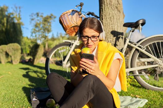 Happy relaxing mature woman in headphones with smartphone looking at screen, communication, video call. Middle-aged female sitting on grass in park eating an apple, bike background, rest and leisure