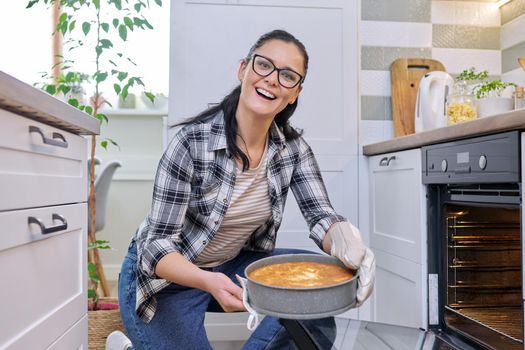 Woman at home in kitchen taking out hot apple pie from oven. Cooking at home, delicious pastries, lifestyle, hobbies and leisure