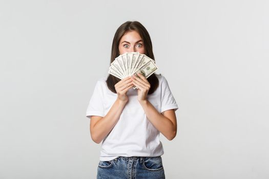 Excited pretty young woman holding money over face, standing white background.
